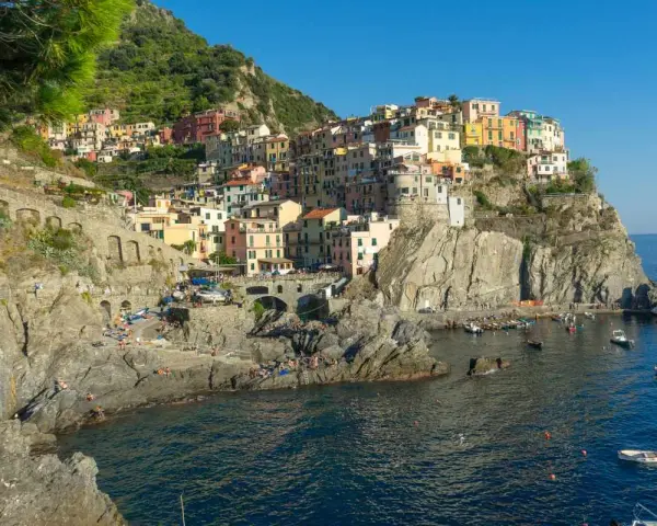 Le Cinque Terre, con le loro case colorate aggrappate alle scogliere, si affacciano sul mare blu. Gente prende il sole sulle rocce.