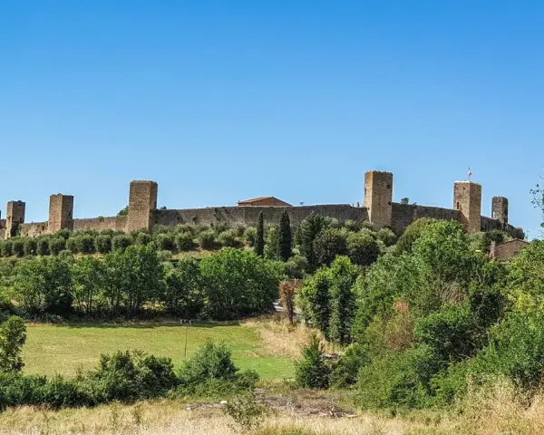 Monteriggioni cosa vedere: imponenti mura medievali con torri quadrate su una collina verdeggiante, sotto un cielo azzurro.