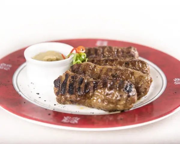 Mici grigliati su un piatto rosso, un classico per sapere cosa mangiare a Bucarest. Serviti con salsa.