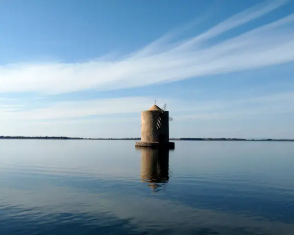 Mulino solitario nella laguna di Orbetello, riflesso sull'acqua calma sotto un cielo azzurro con nuvole sottili.