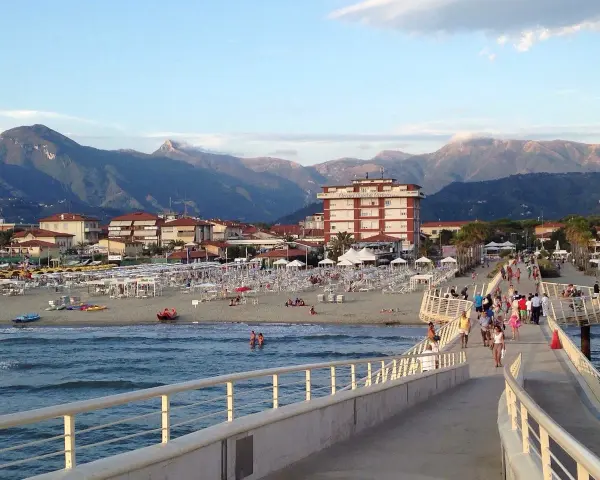 Passeggiata sul molo di Camaiore, dove si trova una spiaggia attrezzata con ombrelloni e montagne sullo sfondo.