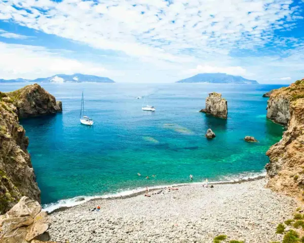 Rocky coastline with a pebble beach, turquoise water, sailboats, and distant islands under a blue, cloudy sky.