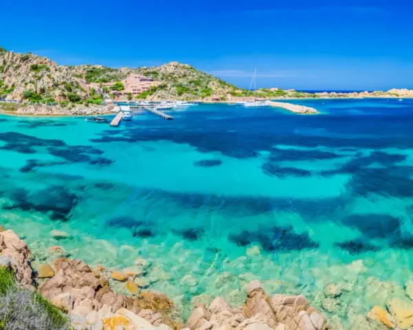 Acque cristalline e rocce granitiche sull'isola della Maddalena. Un porto con yacht e un hotel si affacciano sul mare turchese.