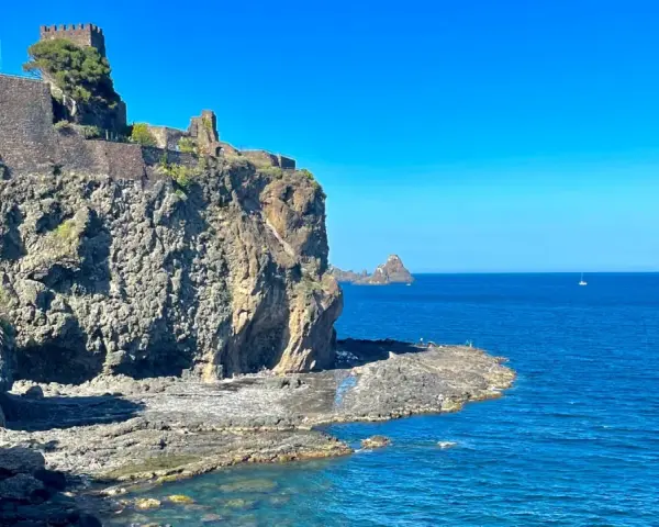 Castello Normanno a Aci Trezza, un luogo da vedere. Roccioso promontorio con antiche mura, mare blu e faraglioni all'orizzonte.