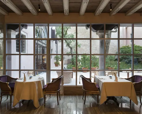 Tavoli apparecchiati in un elegante ristorante stellato Veneto, con vista su un cortile interno con alberi e piante in vaso.