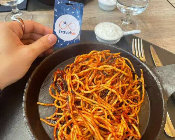 A hand holds a Travel365 tag over a plate of spaghetti with red sauce.