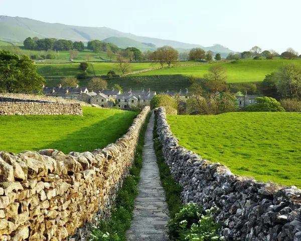 Sentiero tra muretti a secco in Yorkshire, con case di pietra e dolci colline verdi.