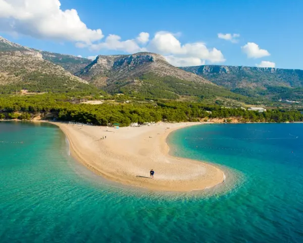 La Croazia offre il mare più bello: una spiaggia dorata a forma di lingua si protende in acque cristalline, con montagne verdi sullo sfondo.