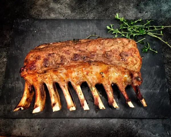 Carré d'agnello arrosto, uno dei 5 piatti tipici della Sardegna, guarnito con erbe aromatiche su una pietra scura.