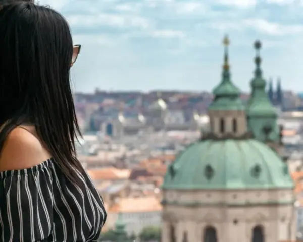 Vista di una donna con capelli scuri appoggiata a un muro di mattoni, con lo sguardo rivolto verso un panorama urbano di **città europee da visitare in 3 giorni**.