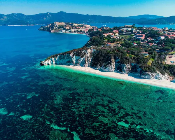 Vista aerea di una spiaggia bianca e mare cristallino, con un borgo arroccato su una collina. Un angolo di paradiso tra le isole arcipelago toscano.