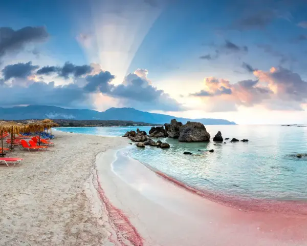 Spiaggia rosa con lettini arancioni e ombrelloni di paglia, mare turchese e montagne sullo sfondo. Un luogo da sogno tra i 10 mari più belli del mondo.