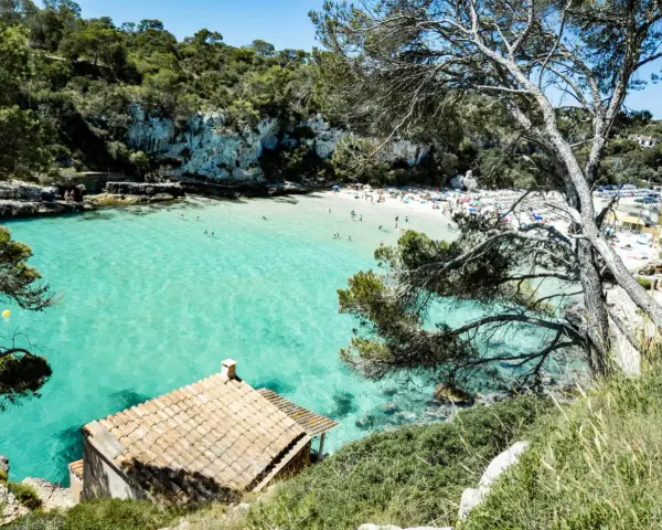 Una baia con acque turchesi cristalline, sabbia bianca e vegetazione lussureggiante. Un piccolo edificio con tetto di tegole si affaccia sul mare. Tra le più belle spiagge di Palma di Maiorca.