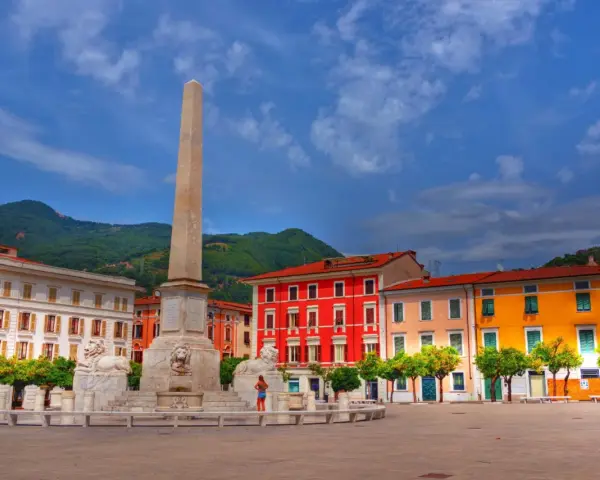 Piazza colorata con obelisco e leoni, incorniciata da edifici vivaci e montagne verdi, nella provincia di Massa e Carrara.