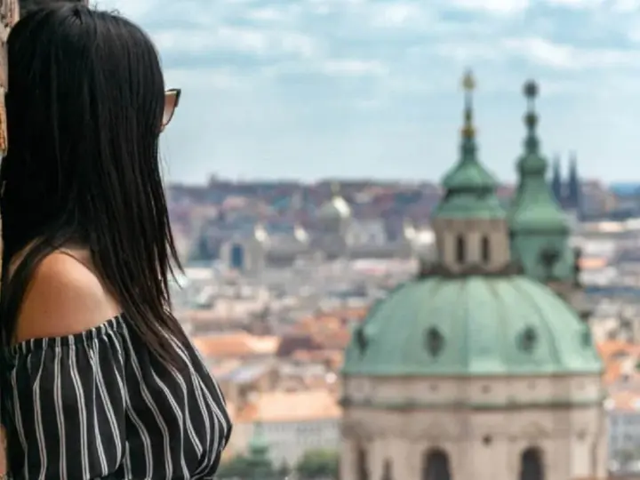 Vista di una donna con capelli scuri appoggiata a un muro di mattoni, con lo sguardo rivolto verso un panorama urbano di **città europee da visitare in 3 giorni**.