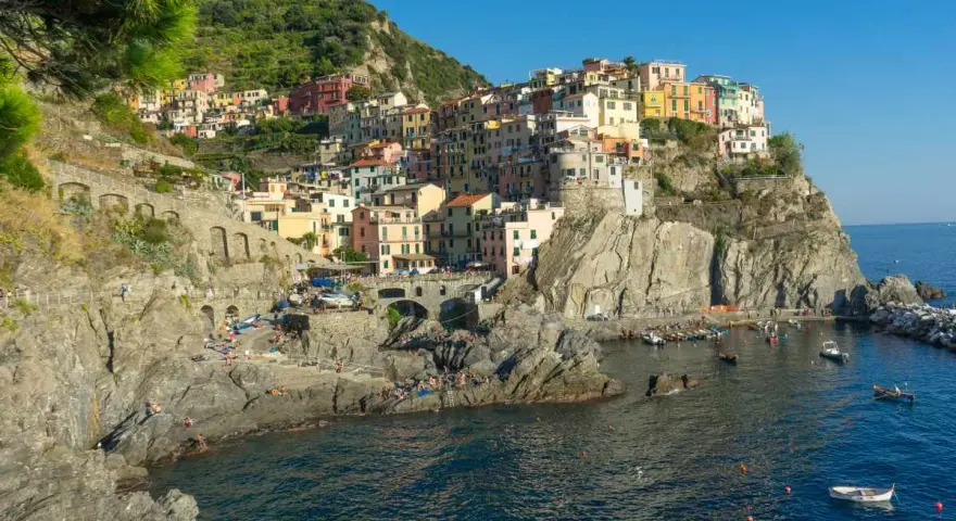 Le Cinque Terre, con le loro case colorate aggrappate alle scogliere, si affacciano sul mare blu. Gente prende il sole sulle rocce.