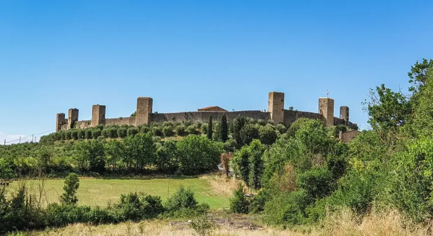 Monteriggioni cosa vedere: imponenti mura medievali con torri quadrate su una collina verdeggiante, sotto un cielo azzurro.