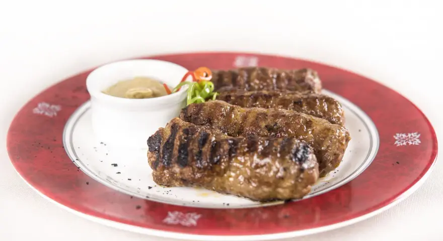 Mici grigliati su un piatto rosso, un classico per sapere cosa mangiare a Bucarest. Serviti con salsa.