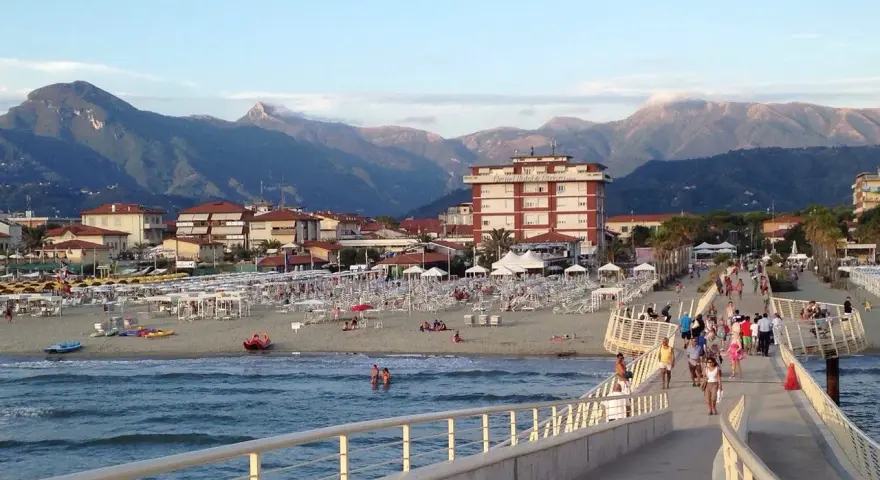 Passeggiata sul molo di Camaiore, dove si trova una spiaggia attrezzata con ombrelloni e montagne sullo sfondo.