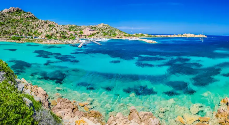 Acque cristalline e rocce granitiche sull'isola della Maddalena. Un porto con yacht e un hotel si affacciano sul mare turchese.