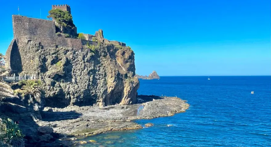 Castello Normanno a Aci Trezza, un luogo da vedere. Roccioso promontorio con antiche mura, mare blu e faraglioni all'orizzonte.