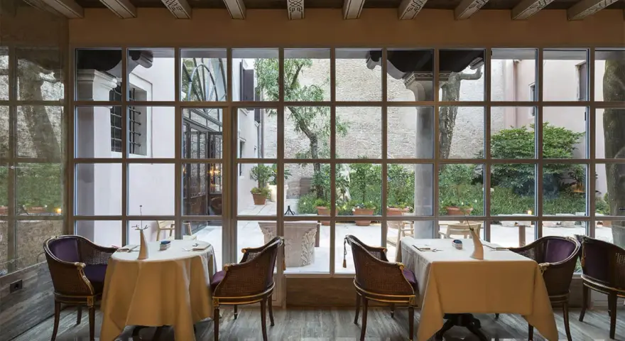 Tavoli apparecchiati in un elegante ristorante stellato Veneto, con vista su un cortile interno con alberi e piante in vaso.