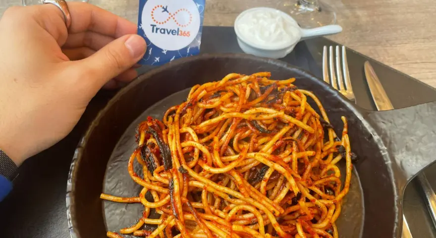 A hand holds a Travel365 tag over a plate of spaghetti with red sauce.