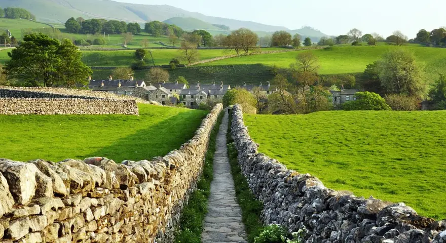 Sentiero tra muretti a secco in Yorkshire, con case di pietra e dolci colline verdi.