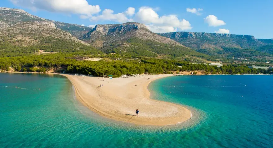 La Croazia offre il mare più bello: una spiaggia dorata a forma di lingua si protende in acque cristalline, con montagne verdi sullo sfondo.