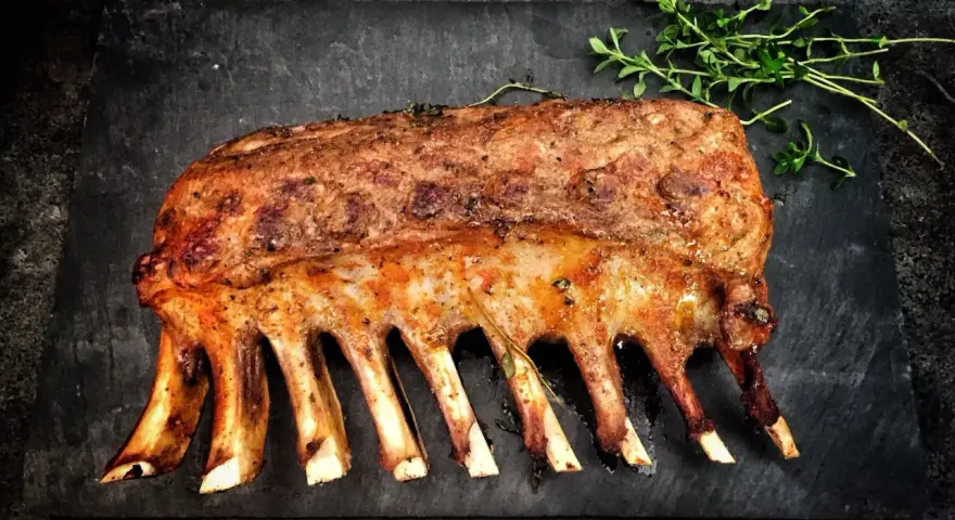 Carré d'agnello arrosto, uno dei 5 piatti tipici della Sardegna, guarnito con erbe aromatiche su una pietra scura.