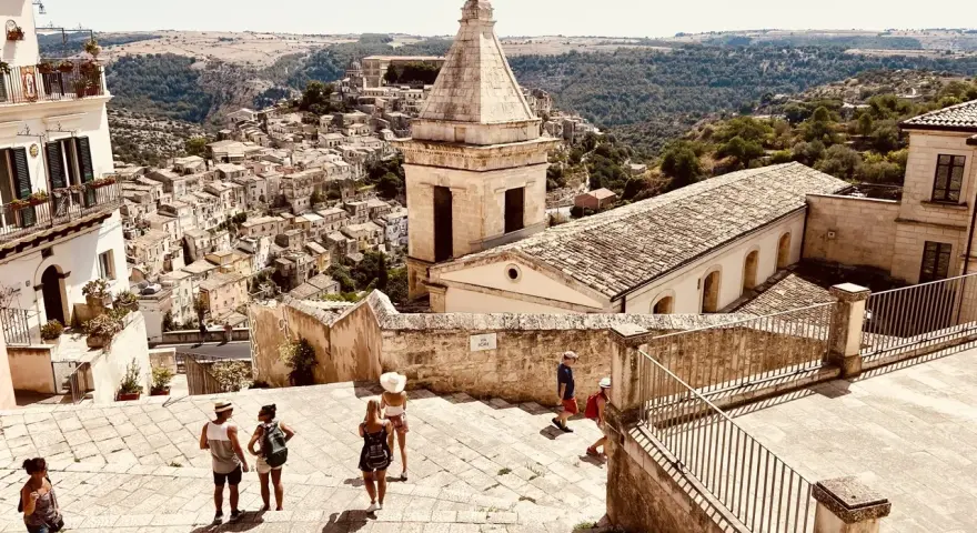 Panoramica di una città barocca in Sicilia orientale, cosa vedere: un borgo arroccato con chiese e case in pietra, turisti che esplorano le scalinate.