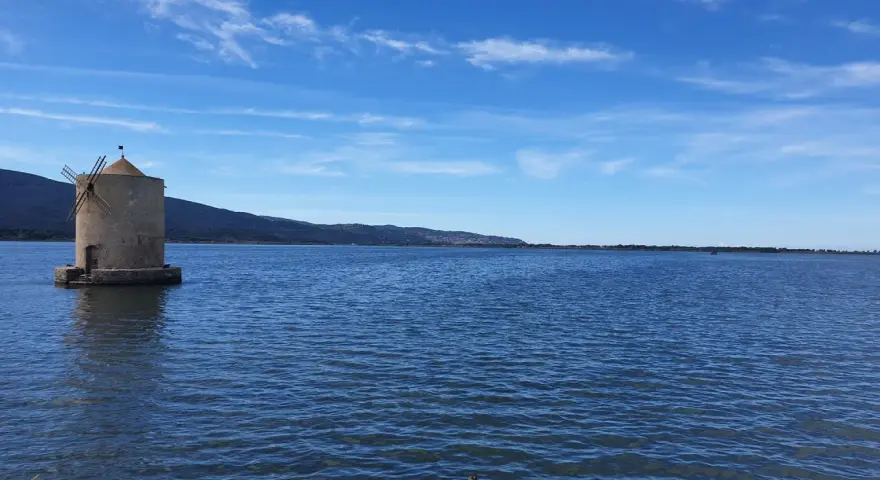 Mulino a Orbetello, un'icona da vedere. Paesaggio lagunare con cielo azzurro e colline sullo sfondo.