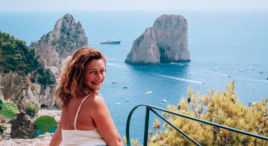 Donna sorridente con vista sui Faraglioni, un'icona dell'isola di Capri. Il mare azzurro e le barche completano il paesaggio.