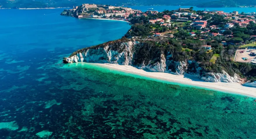 Vista aerea di una spiaggia bianca e mare cristallino, con un borgo arroccato su una collina. Un angolo di paradiso tra le isole arcipelago toscano.