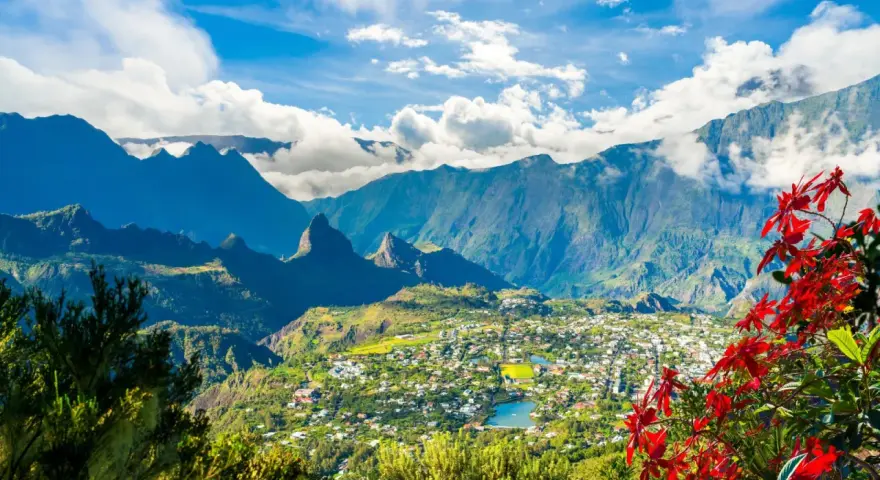Vista panoramica di un villaggio verdeggiante sull'isola di Reunion, incastonato tra imponenti montagne blu e fiori rossi vibranti.
