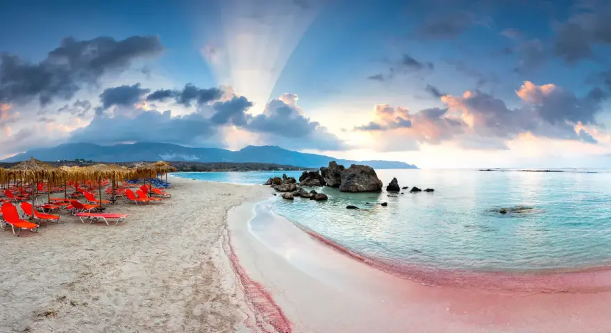Spiaggia rosa con lettini arancioni e ombrelloni di paglia, mare turchese e montagne sullo sfondo. Un luogo da sogno tra i 10 mari più belli del mondo.