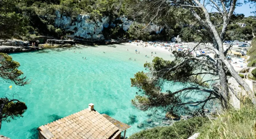 Una baia con acque turchesi cristalline, sabbia bianca e vegetazione lussureggiante. Un piccolo edificio con tetto di tegole si affaccia sul mare. Tra le più belle spiagge di Palma di Maiorca.
