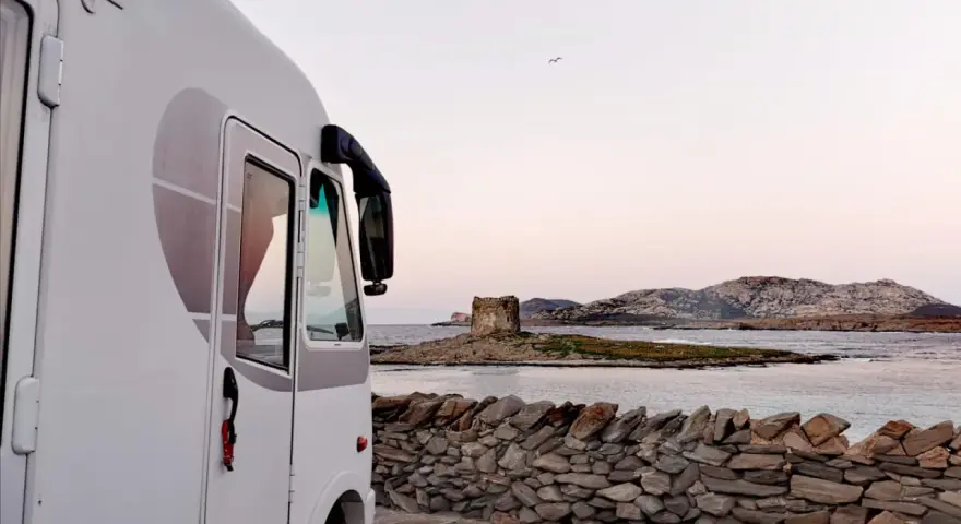 Un camper bianco parcheggiato su un lastricato di pietra, con una vista mozzafiato sul mare e una torre antica in lontananza. Un sogno di **Sardegna in camper**.