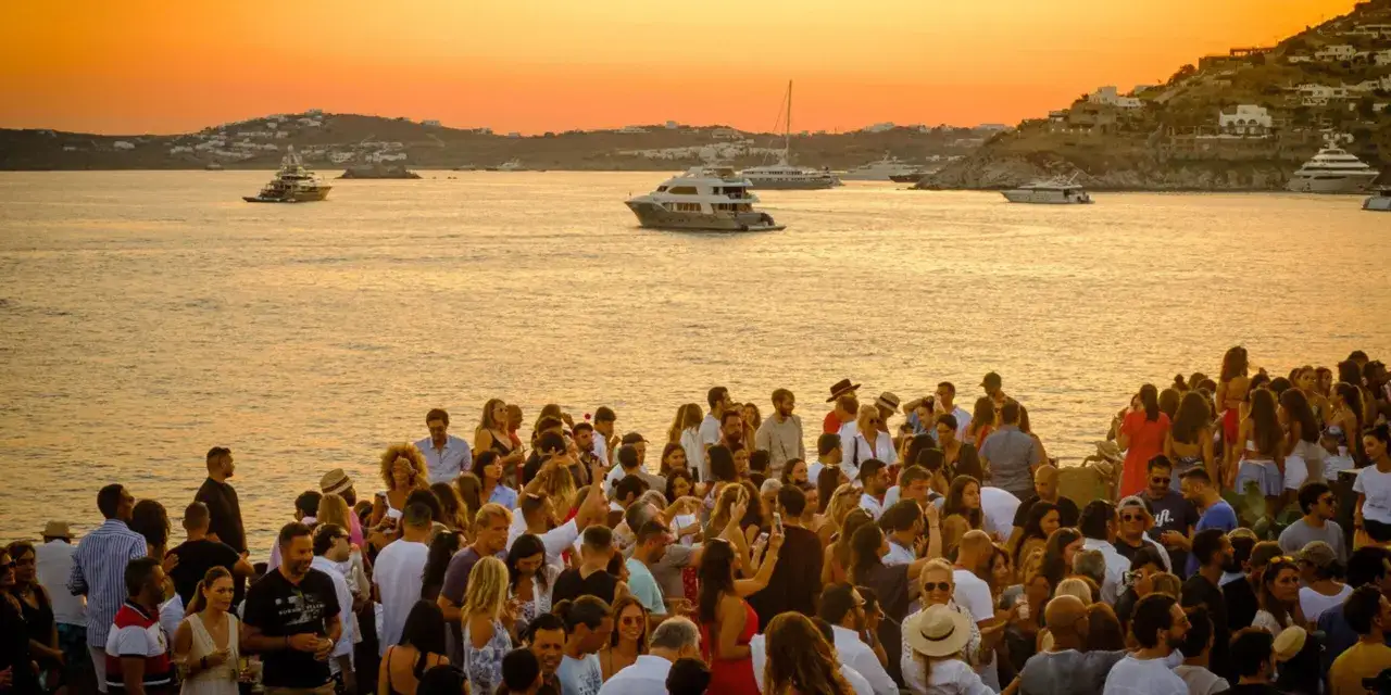 Festa al tramonto su una delle isole greche per giovani, con yacht all'orizzonte e gente che si diverte sulla costa.