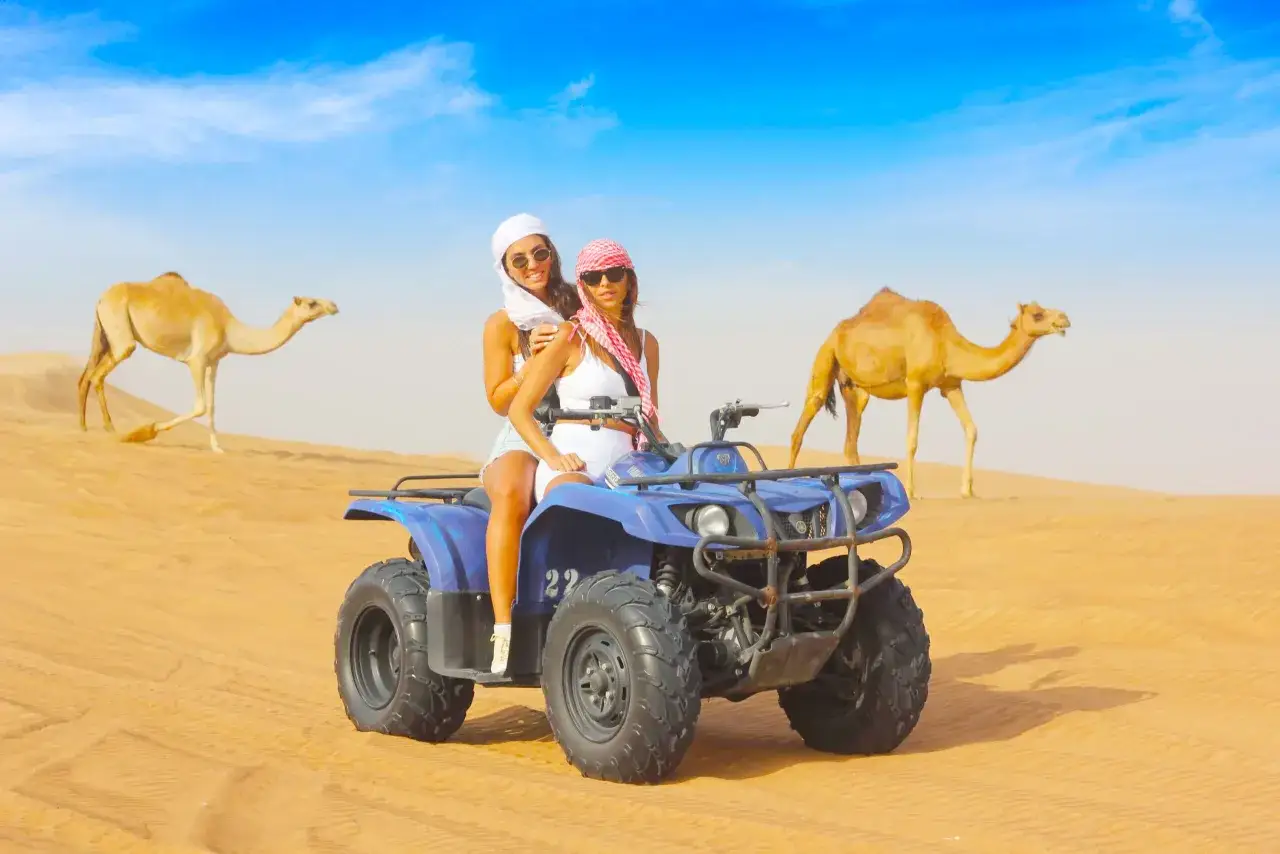 Due donne su un quad nel deserto, con cammelli sullo sfondo. Un'avventura indimenticabile per le escursioni a Marsa Alam.