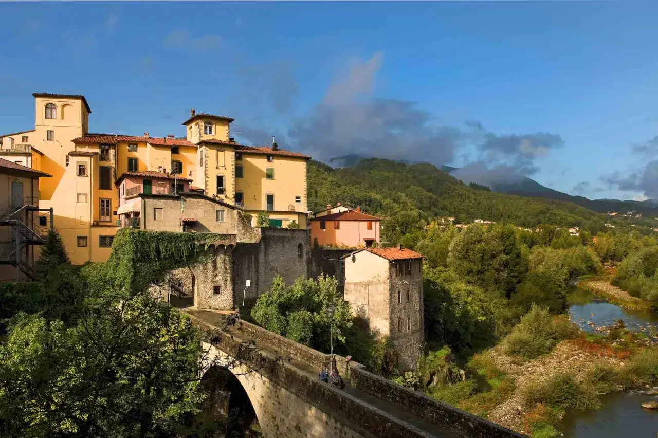 Ponte medievale a Castelnuovo di Garfagnana, con case colorate arroccate sulla collina e un fiume che scorre tra gli alberi.