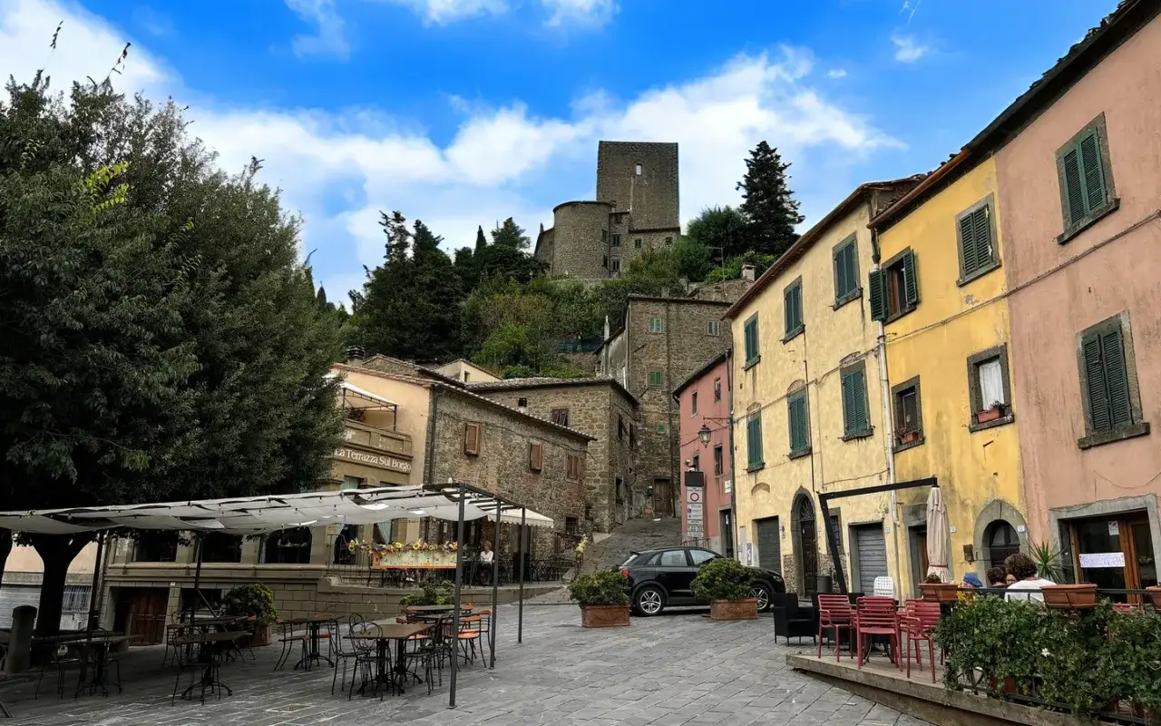 Piazza nel Val di Cecina con case colorate, un castello in collina e tavolini all'aperto.