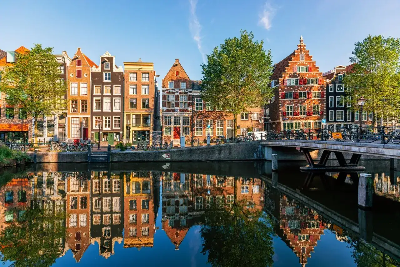 Case di Amsterdam lungo un canale, perfette per scoprire le citt&agrave; europee da visitare in 3 giorni. Riflessi sull'acqua e biciclette ovunque.