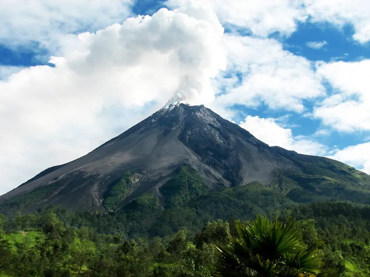Fumo sale dal vulcano attivo sull'isola di Reunion, circondato da lussureggiante vegetazione verde sotto un cielo azzurro con nuvole bianche.