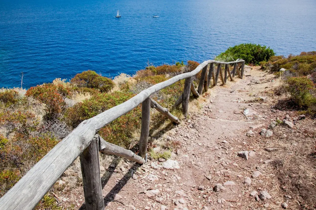 Sentiero con staccionata in legno sull'isola di Capraia, con il mare blu e due barche a vela in lontananza.
