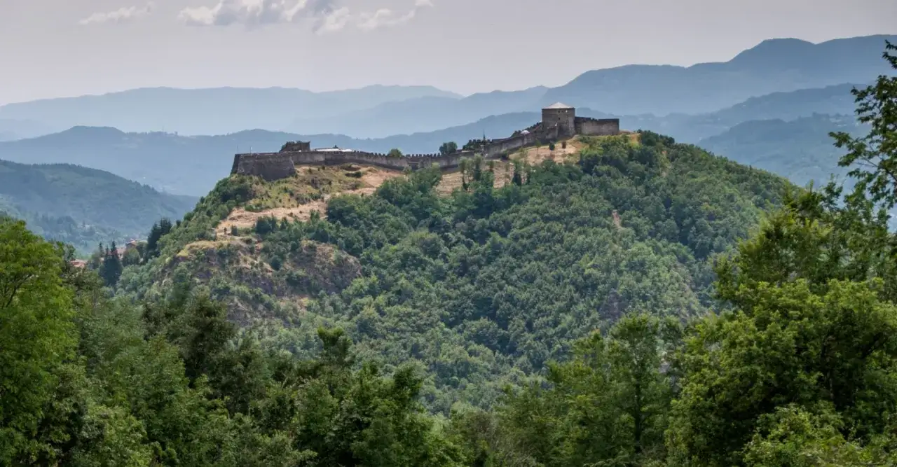 Rocche imponenti dominano la collina boscosa di Castelnuovo di Garfagnana, con le montagne azzurre che si stagliano all'orizzonte.