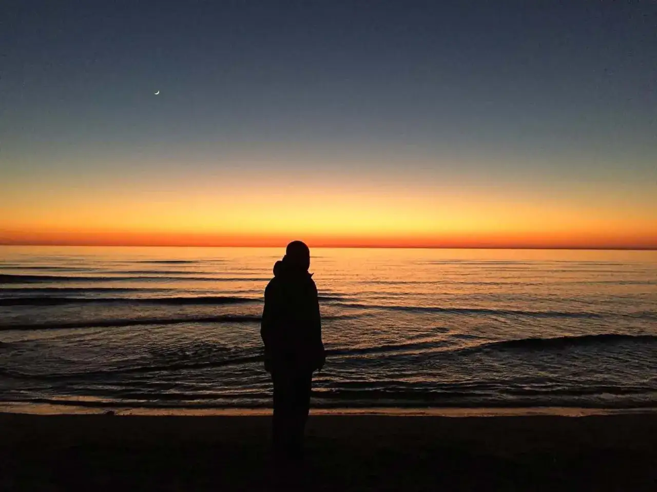 Silhouette di una persona ammira il tramonto sul mare al Lido di Camaiore, con la luna crescente nel cielo.