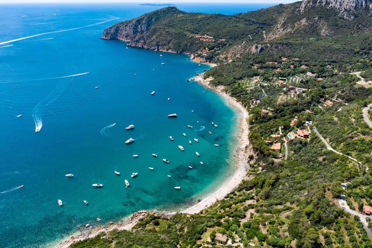 Baia con acque cristalline e numerose barche, incastonata nella costa verdeggiante del Monte Argentario, dove si trova questo splendido tratto di mare.