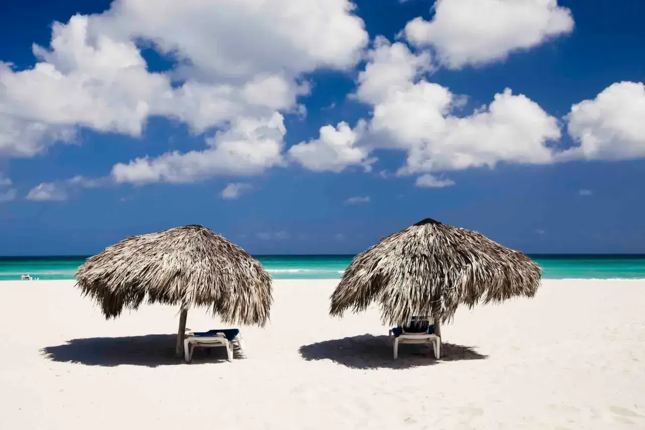 Due lettini vuoti sotto ombrelloni di paglia su una spiaggia bianca, con il mare turchese e nuvole bianche nel cielo azzurro. Ecco cosa vedere a Cuba per una vacanza da sogno.