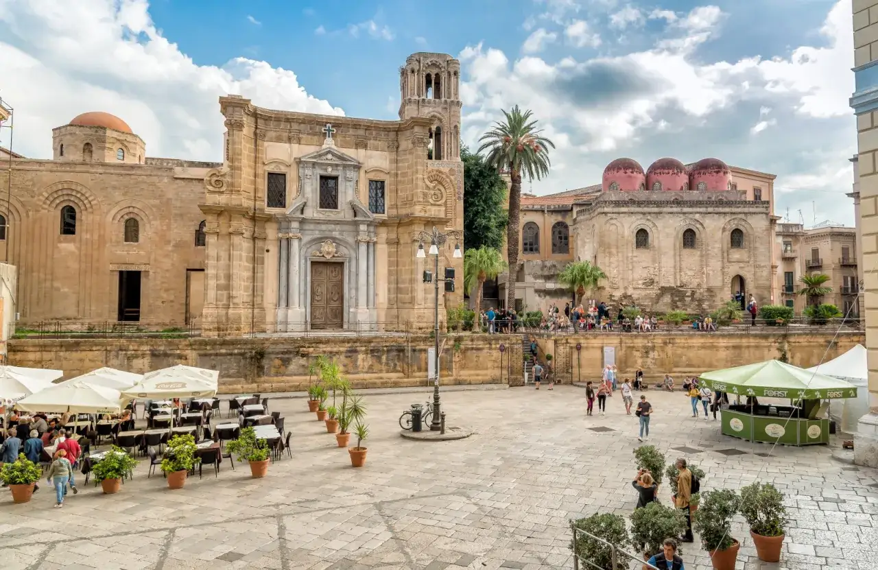 Piazza con chiese normanne, un luogo da scoprire tra le cose da vedere a Palermo. Tavolini all'aperto e gente passeggia.